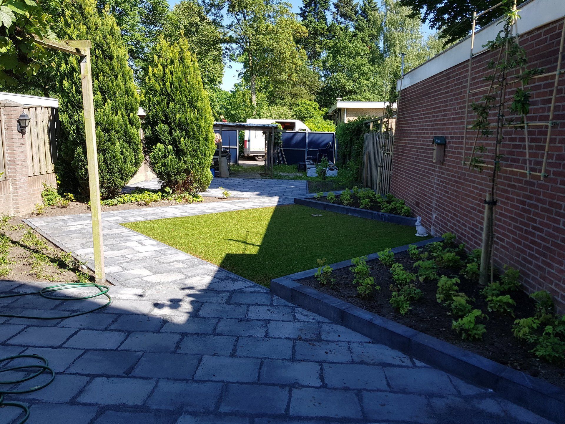 Terras in de achtertuin met stenen bestrating, groen gazon, struiken en een bakstenen huis in het volle zonlicht.