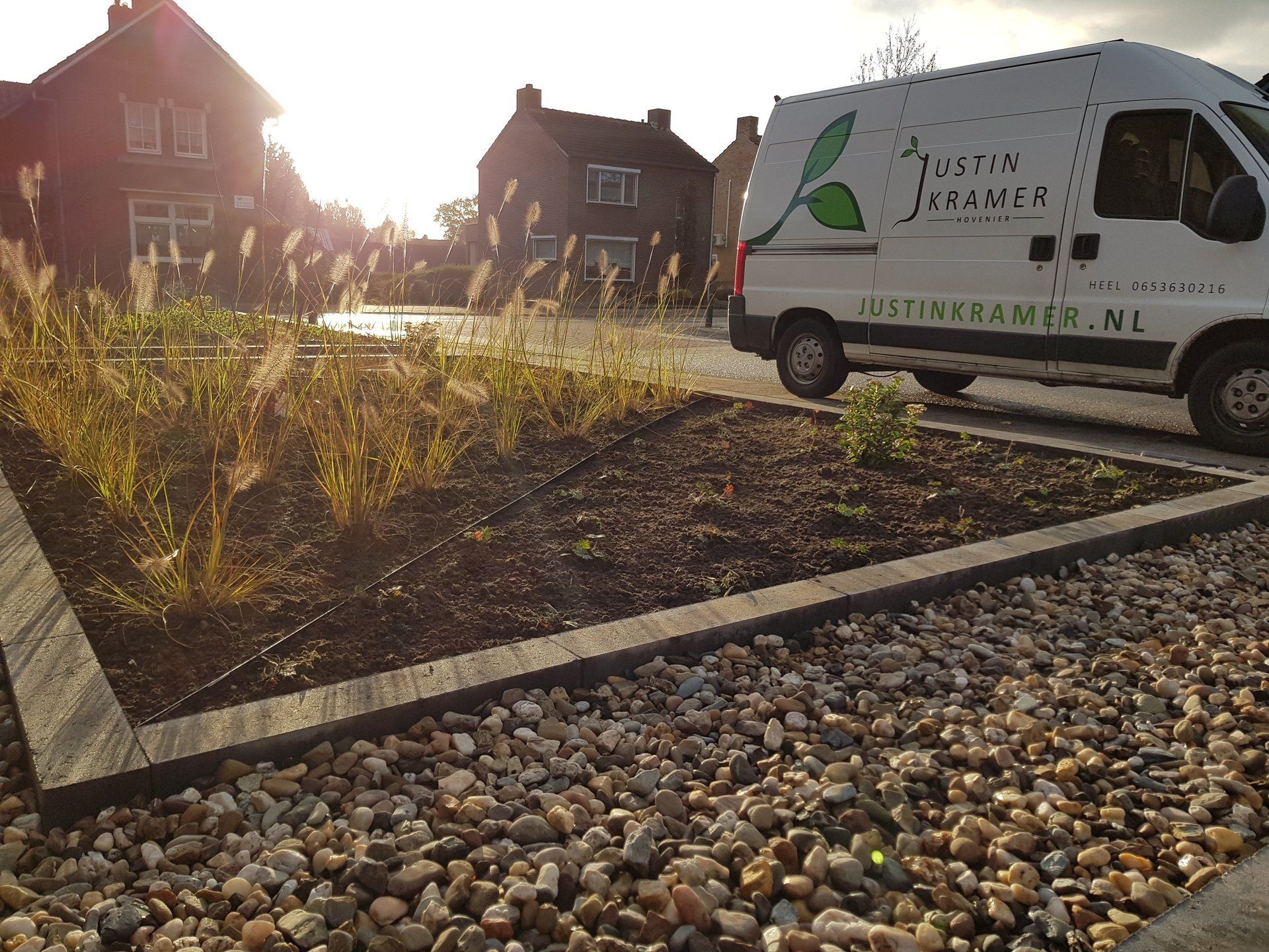 Een witte servicebus staat geparkeerd naast een grindpad en een aangelegde tuin bij zonsondergang.