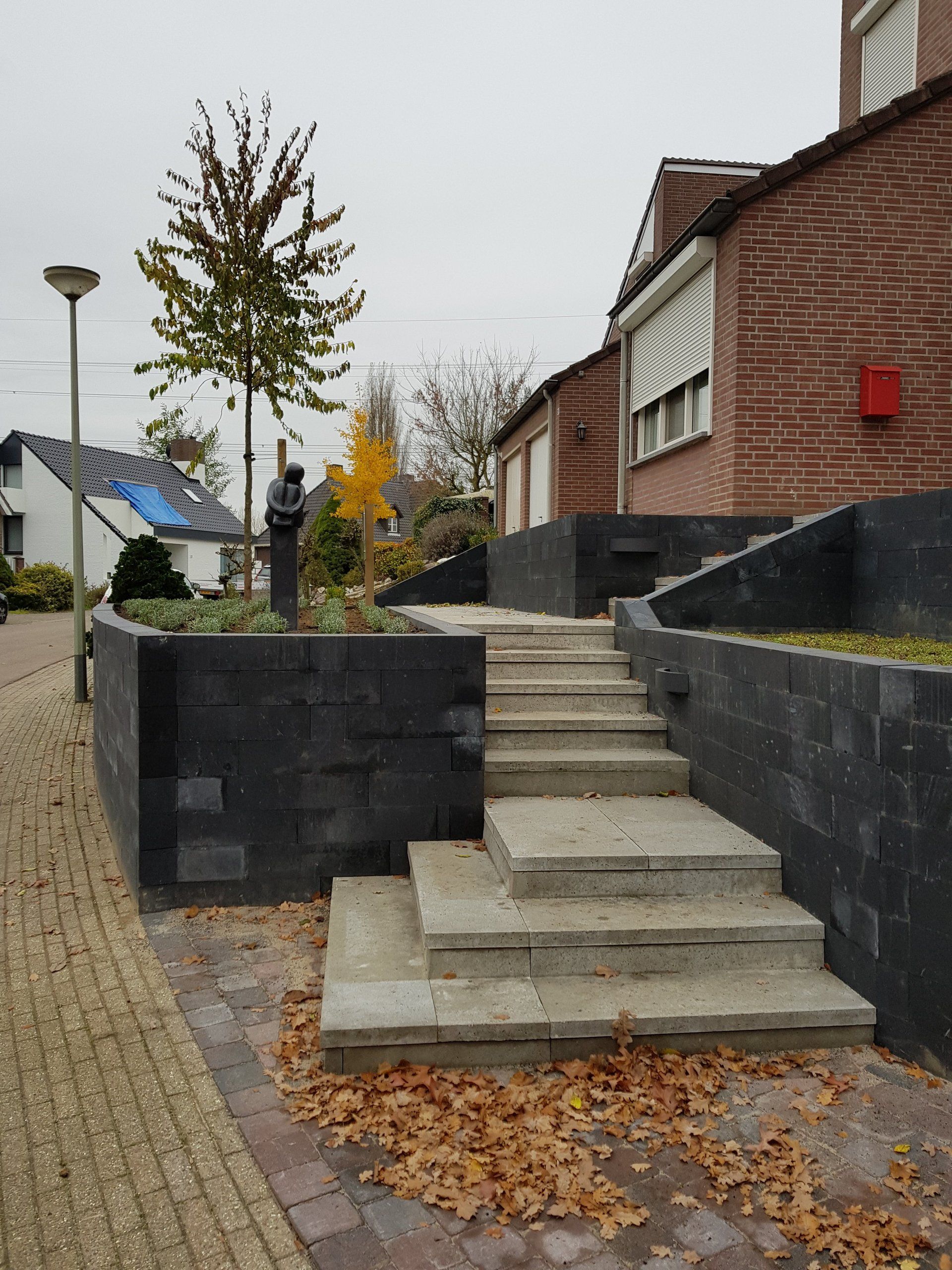 Buitentrap met donkere bakstenen keermuren naast een trottoir en een gebouw, bedekt met gevallen bladeren.