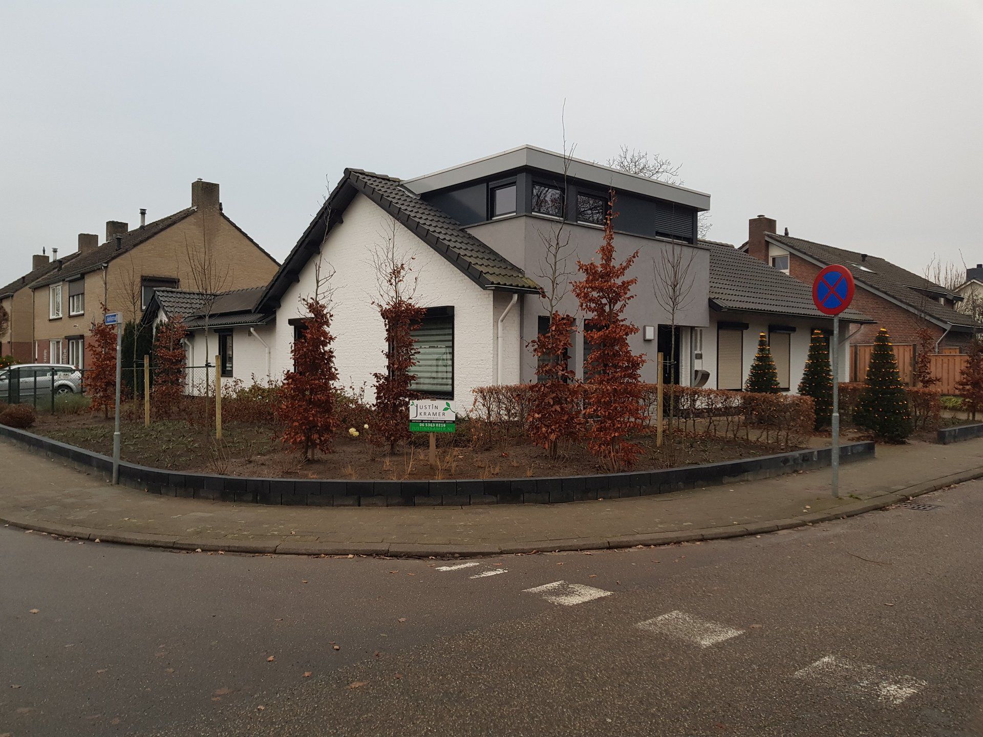Straathoek met een wit huis, struiken met rode bladeren en een parkeerverbodsbord onder een bewolkte hemel.