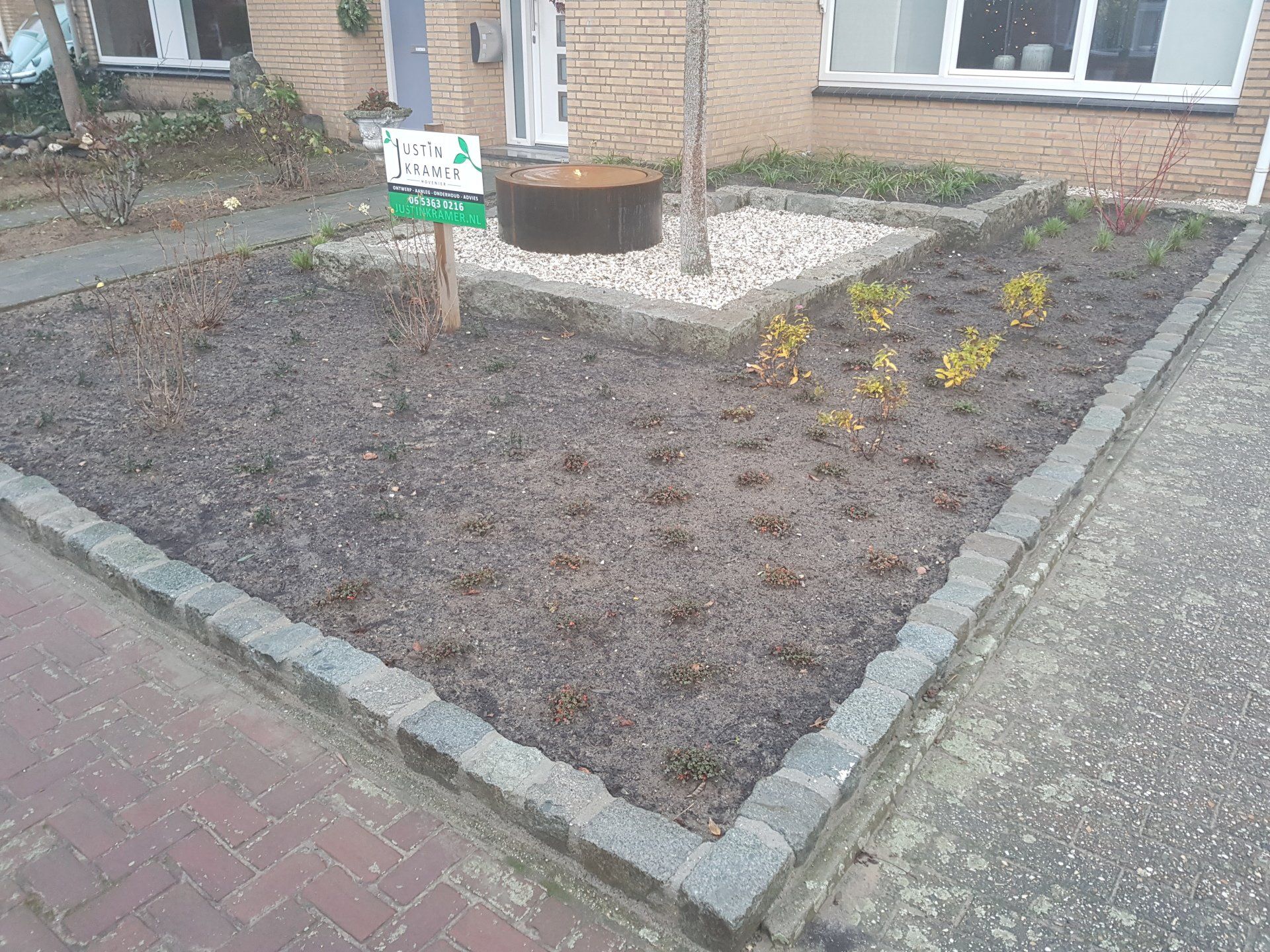Een pas aangelegde voortuin met mulch, stenen rand en een klein bordje naast een bakstenen huis.