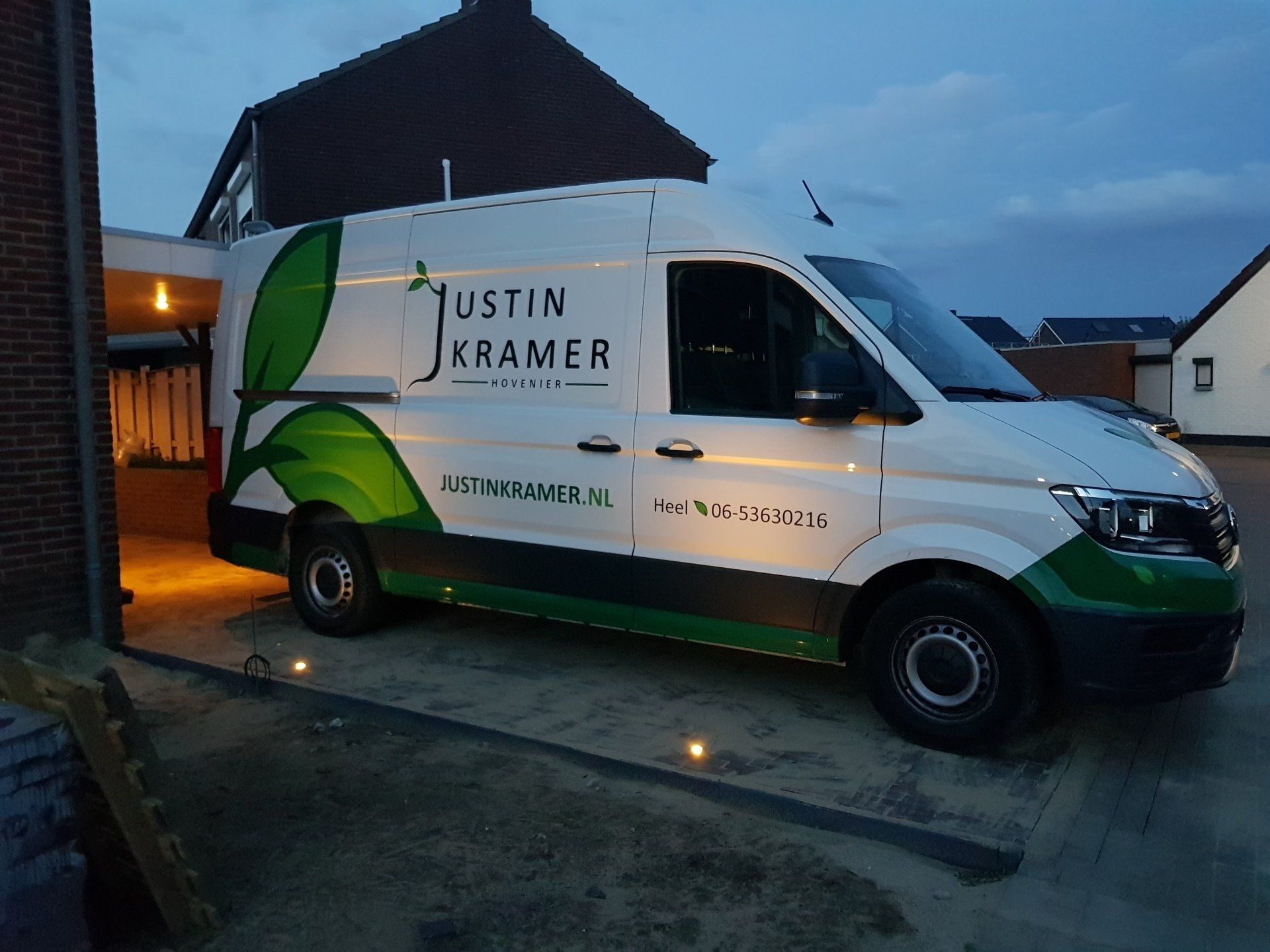 Een wit-groene Justin Kramer-bestelbus staat geparkeerd voor een huis in de schemering.
