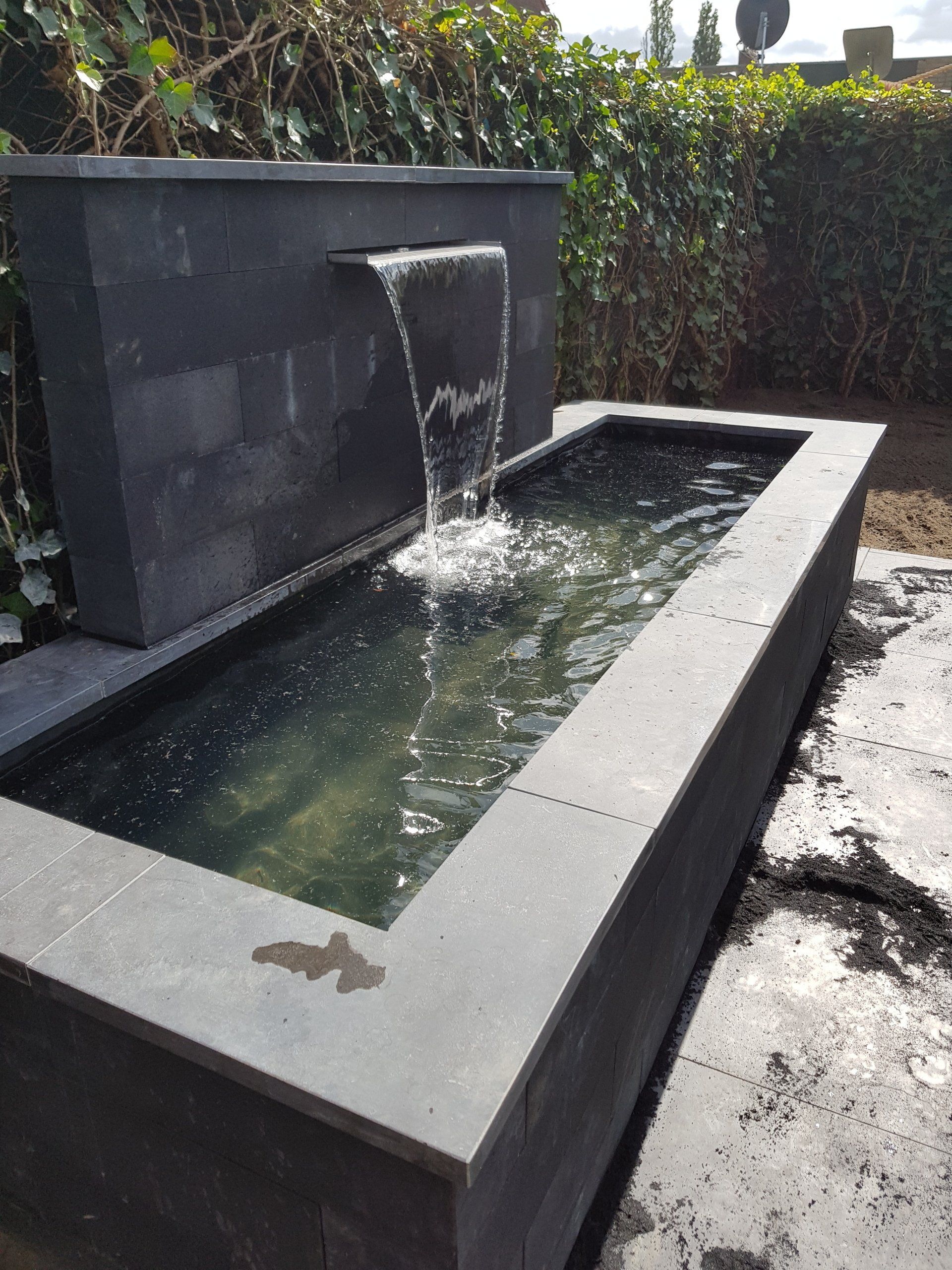 Moderne fontein van zwart steen met water dat in een rechthoekig reflecterend bassin buiten stroomt.