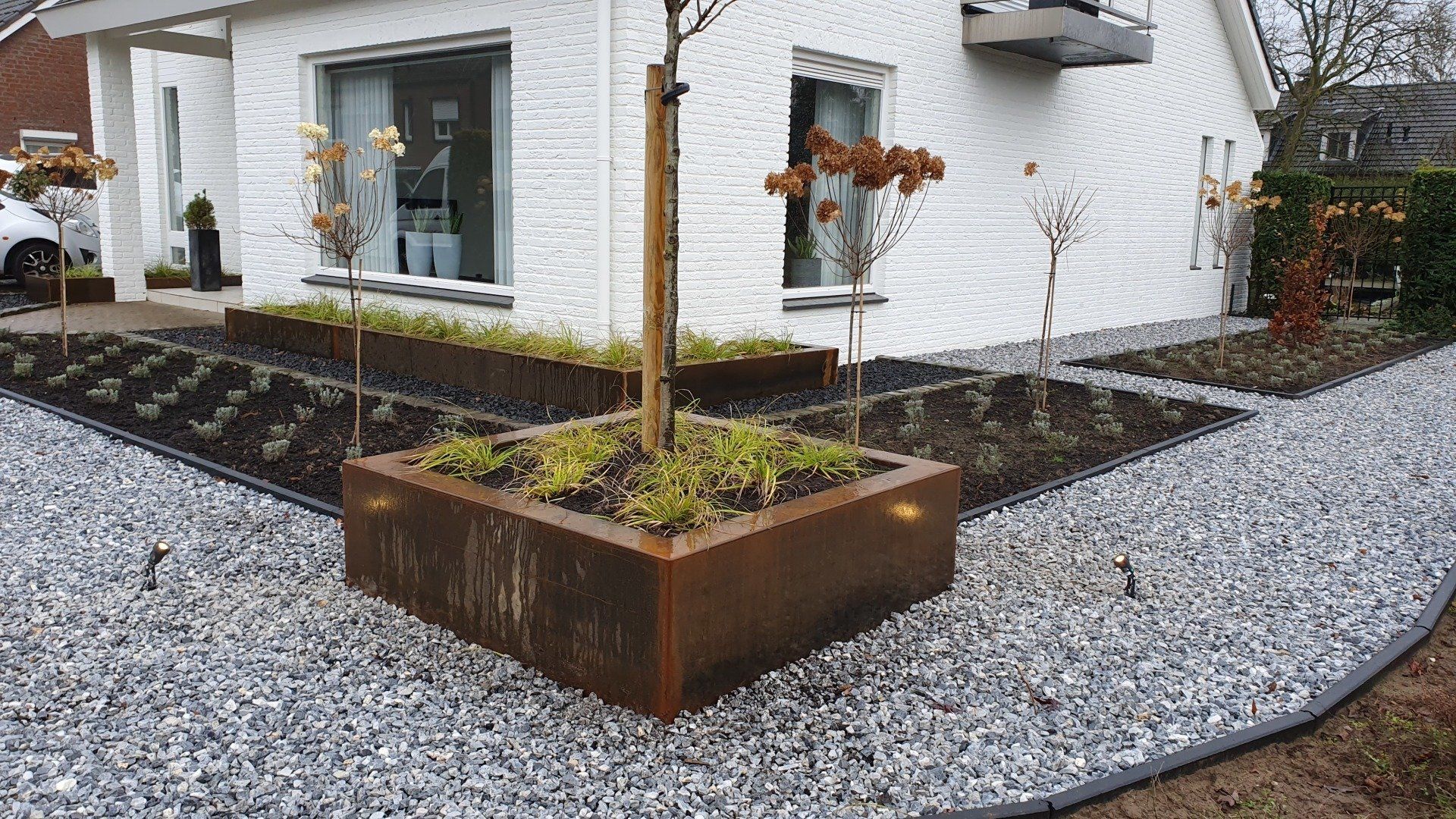Moderne huisingang met grindtuin en een roestkleurige plantenbak rond een jonge boom.