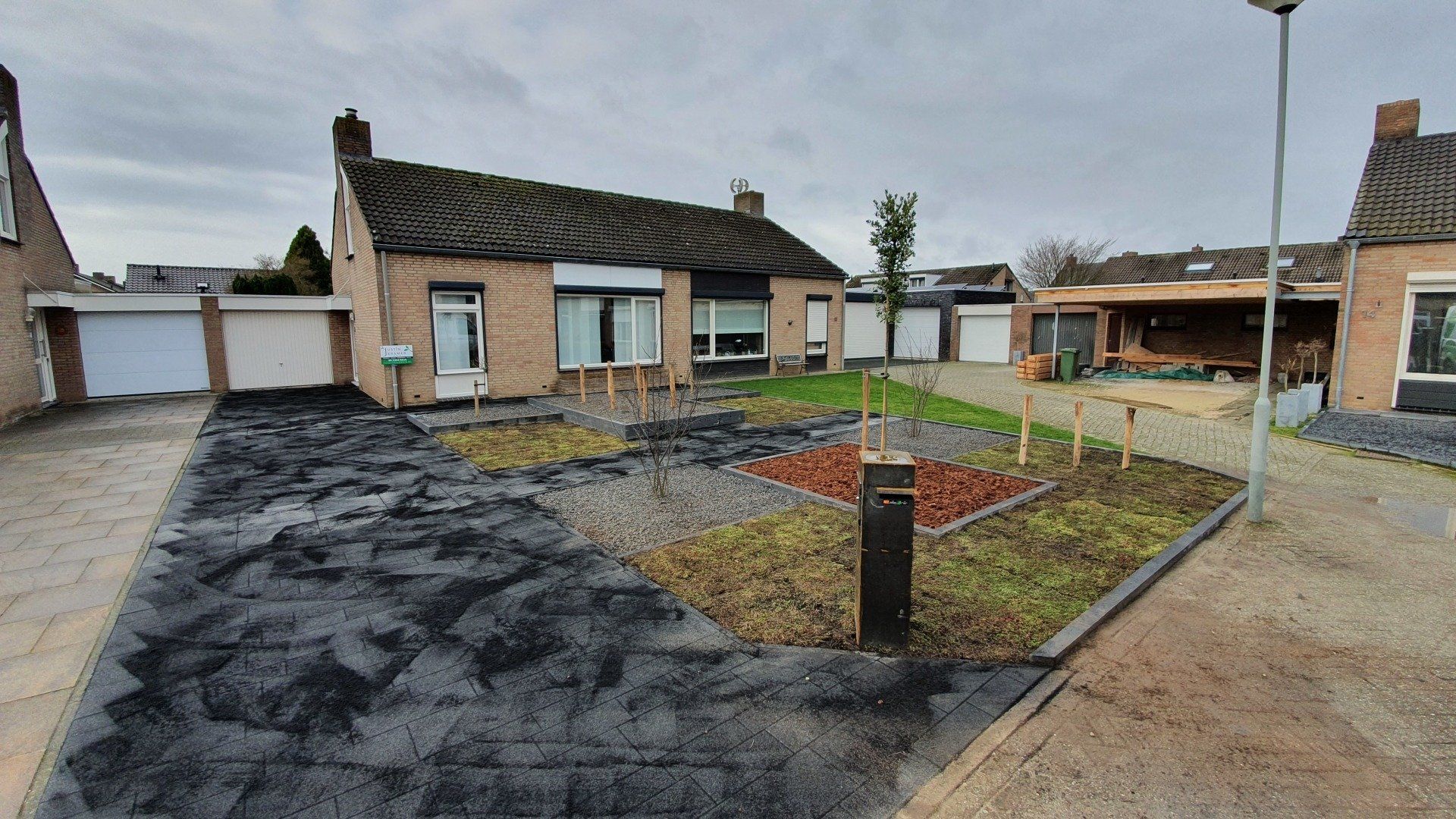 Beschadigde oprit en voortuin van een eengezinswoning die wordt gerenoveerd, met kale grond en bestratingswerkzaamheden in uitvoering.