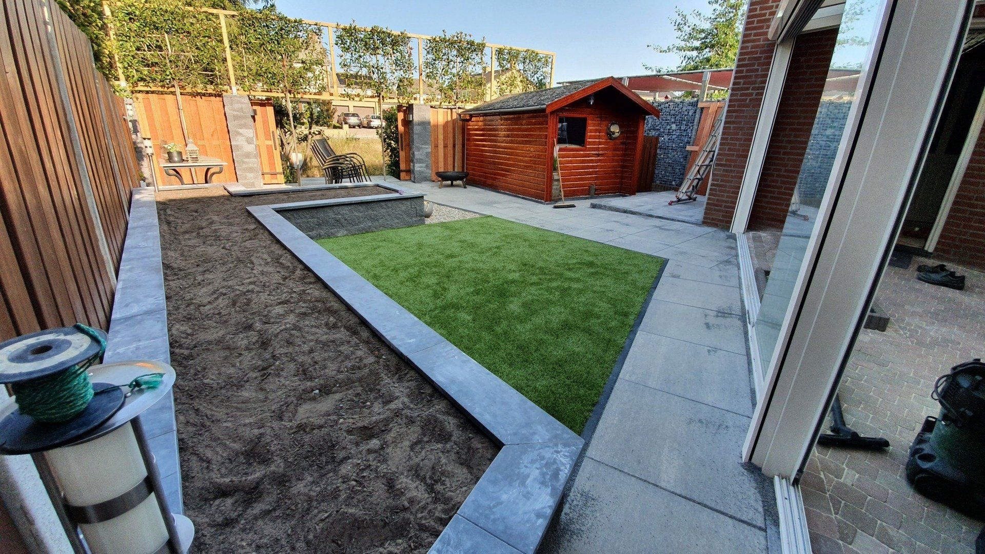 Terras in de achtertuin met een centrale strook gazon, bestrating, schuur en schutting, zichtbaar vanuit een deuropening.