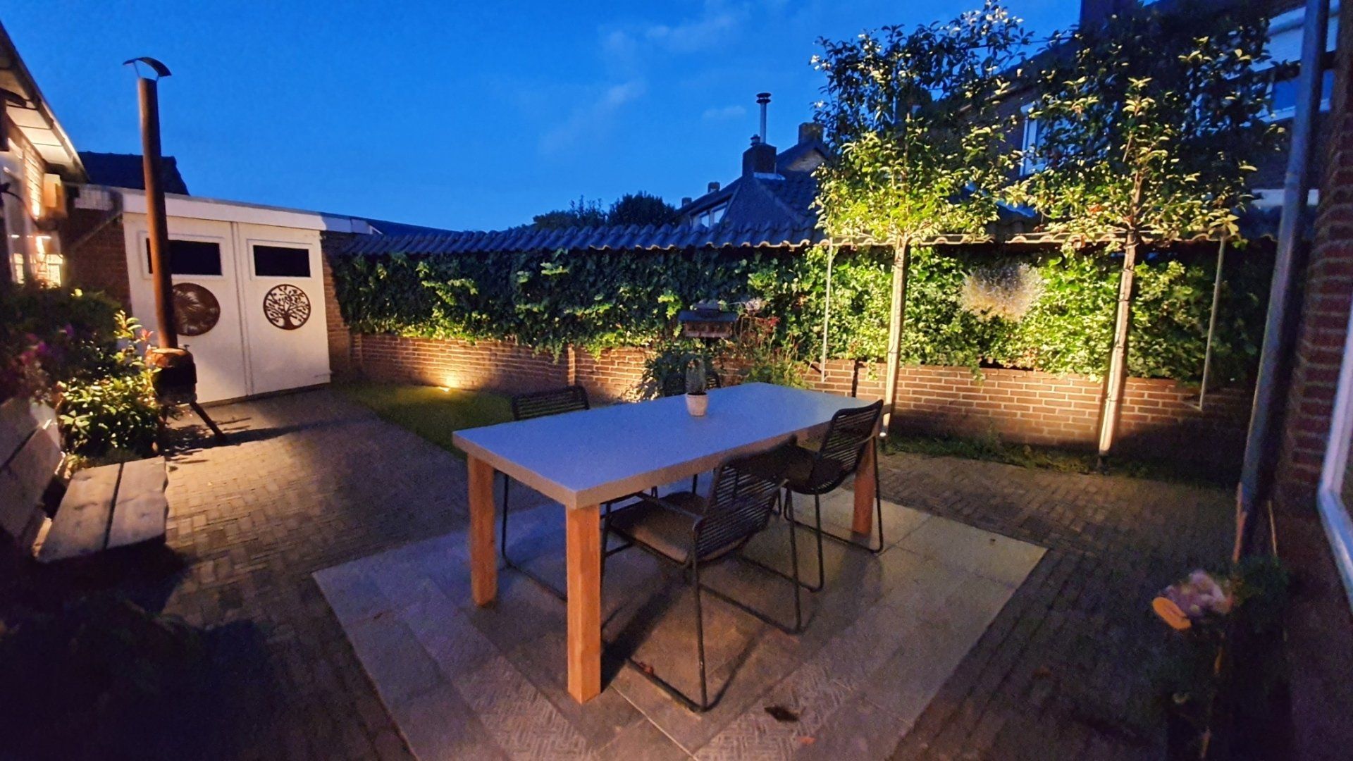 Een terras in de schemering met een houten tafel, stoelen, warme verlichting en omringend groen onder een blauwe hemel.