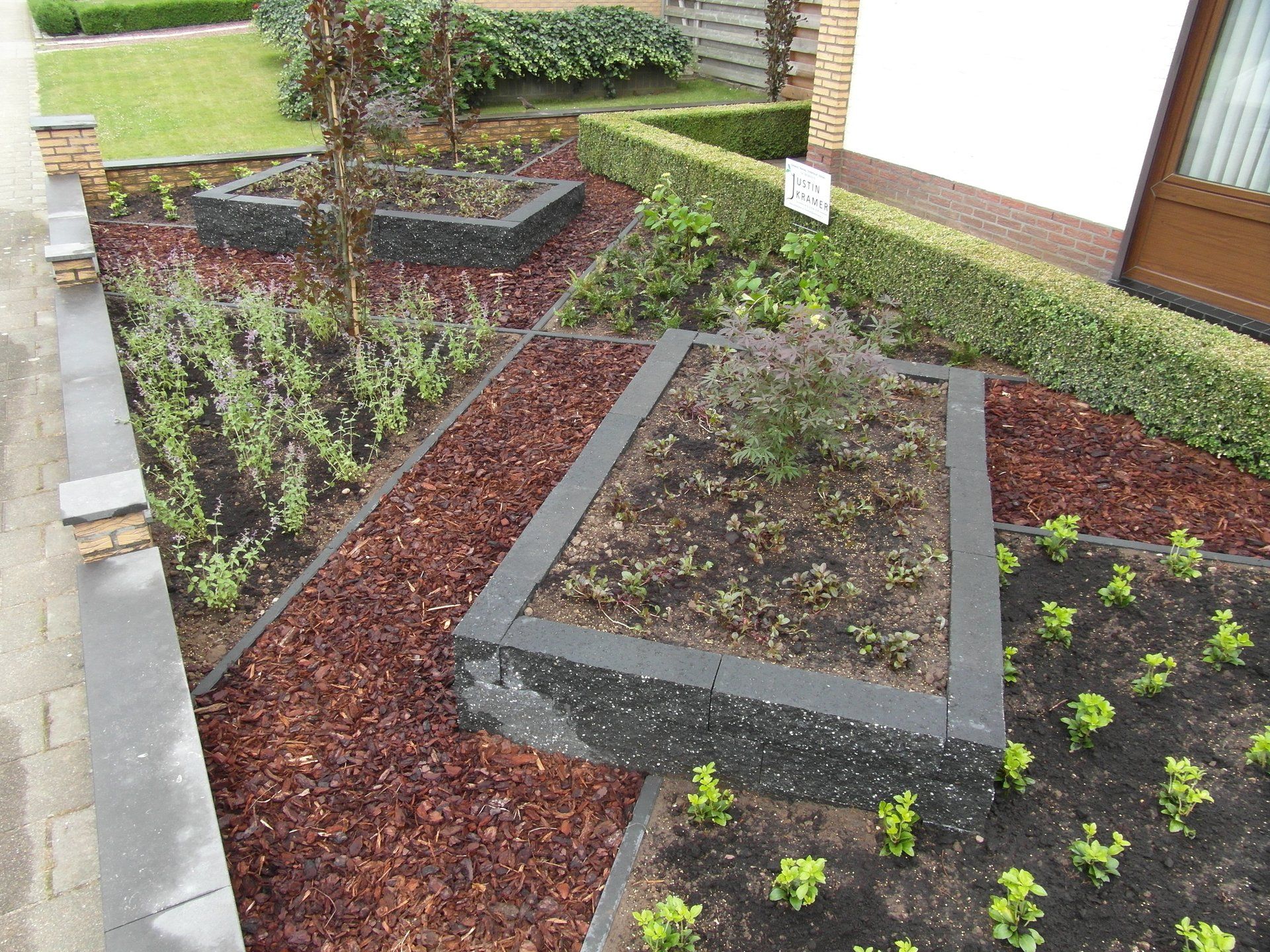 Verhoogde plantenbedden met rode mulch en kleine plantjes naast een looppad langs het huis.