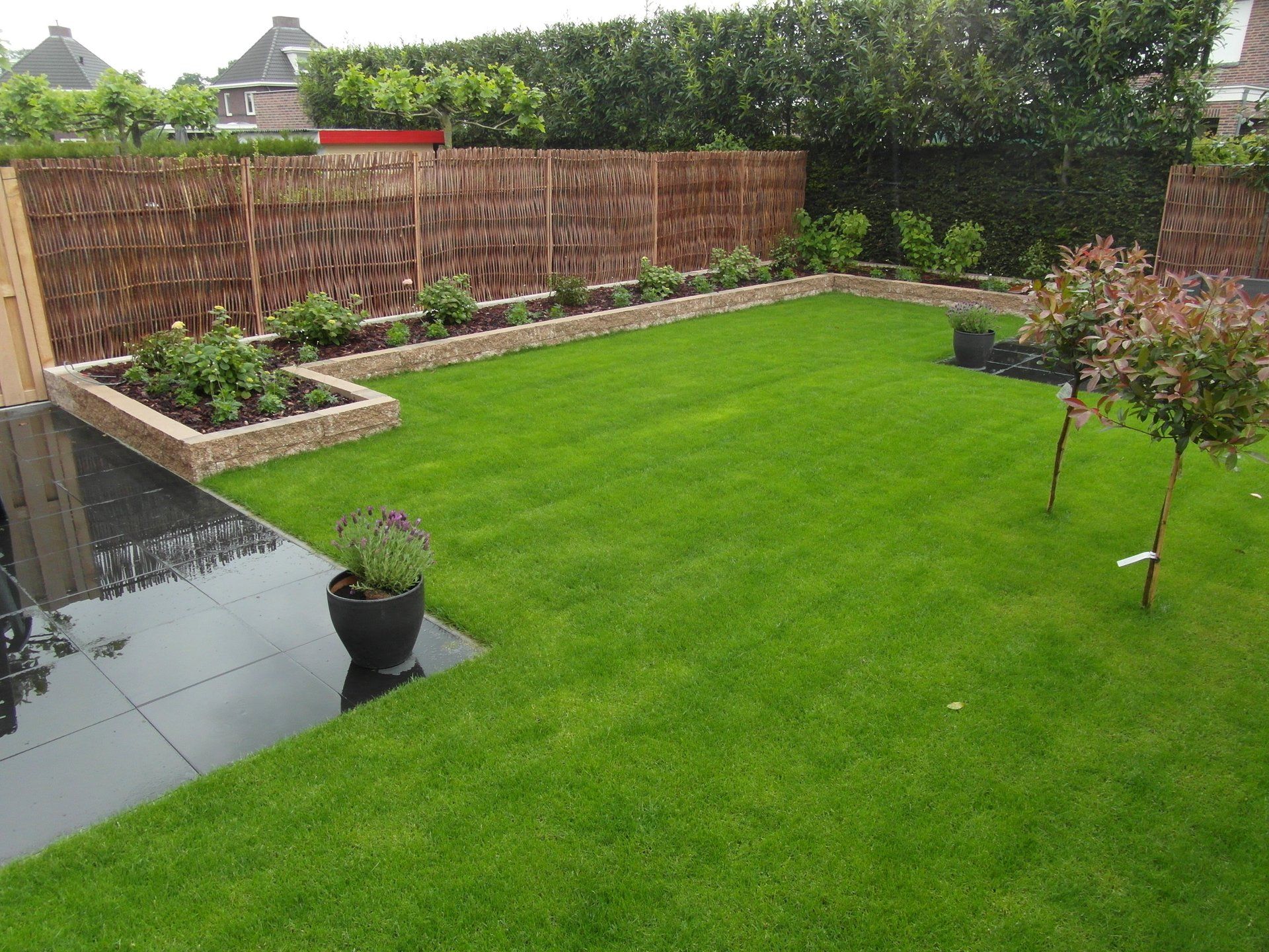 Achtertuin met een weelderig groen gazon, verhoogde bloembedden, een houten schutting en een klein terras aan het water.
