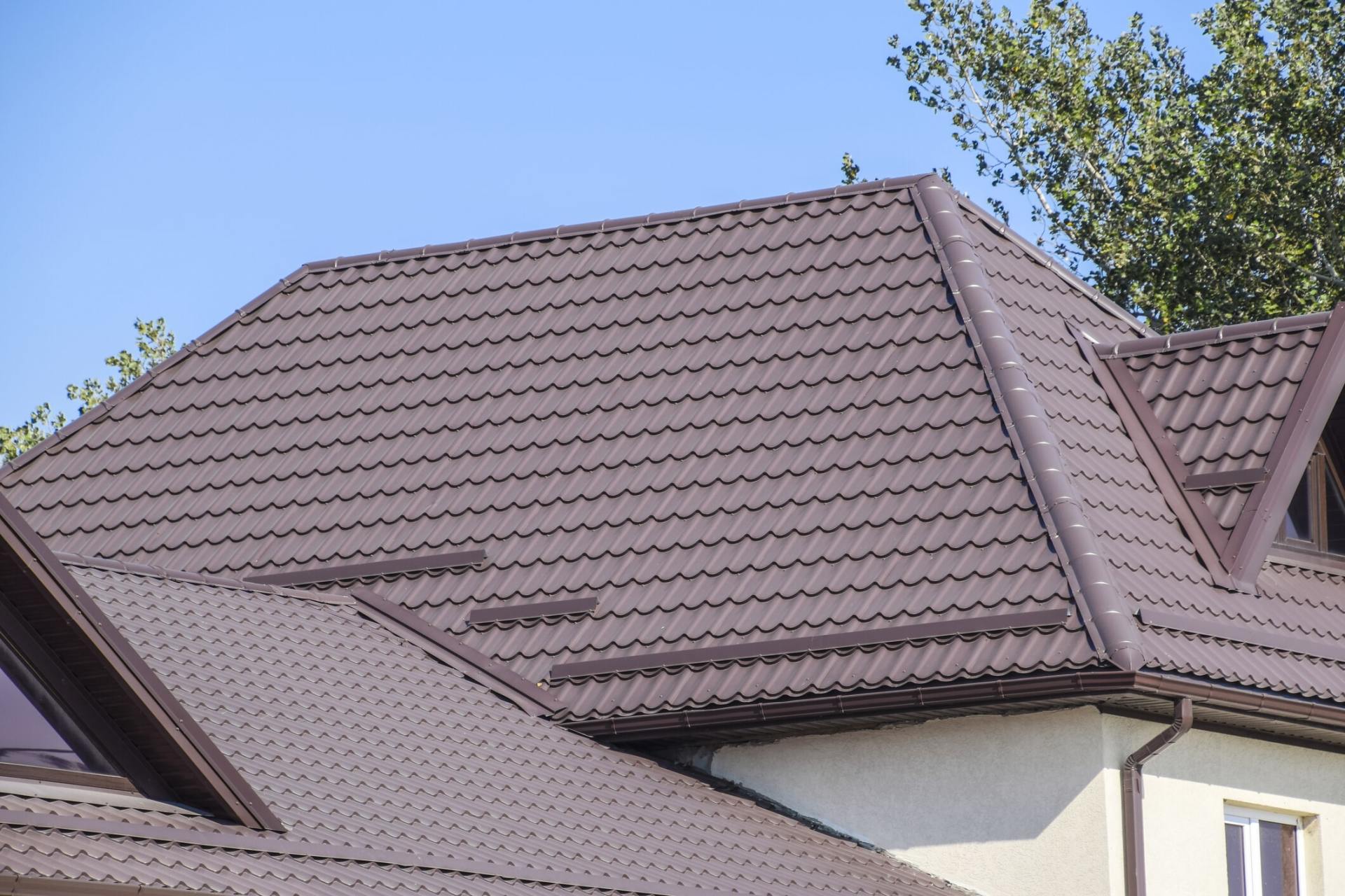 Brown metal roof on a house with dormers against a blue sky and green trees.