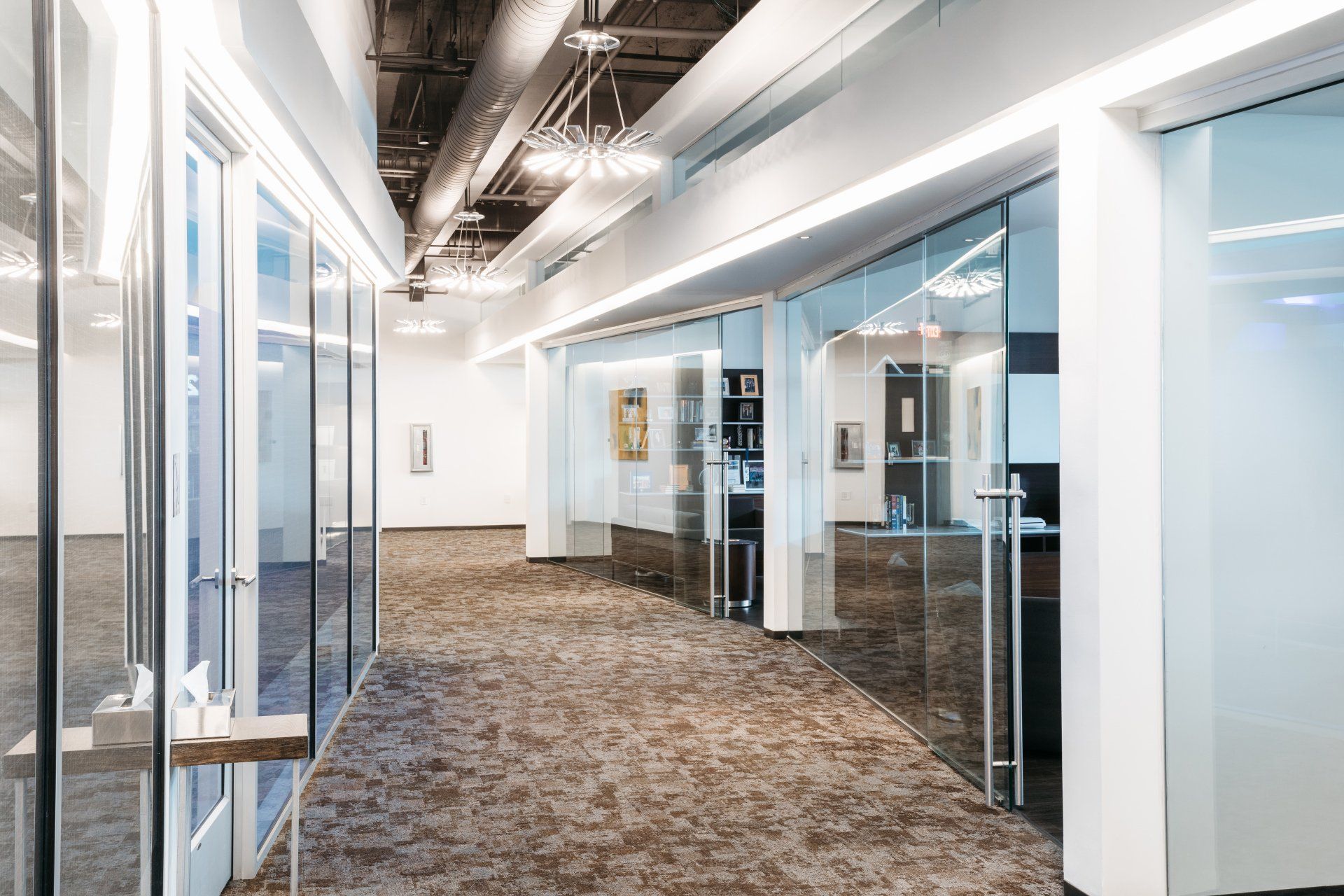Modern glass-walled office hallway with carpeted floor and overhead lighting