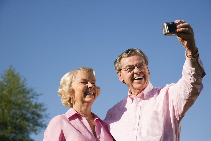 Elderly couple taking a selfie outdoors, both smiling with a blue sky background.