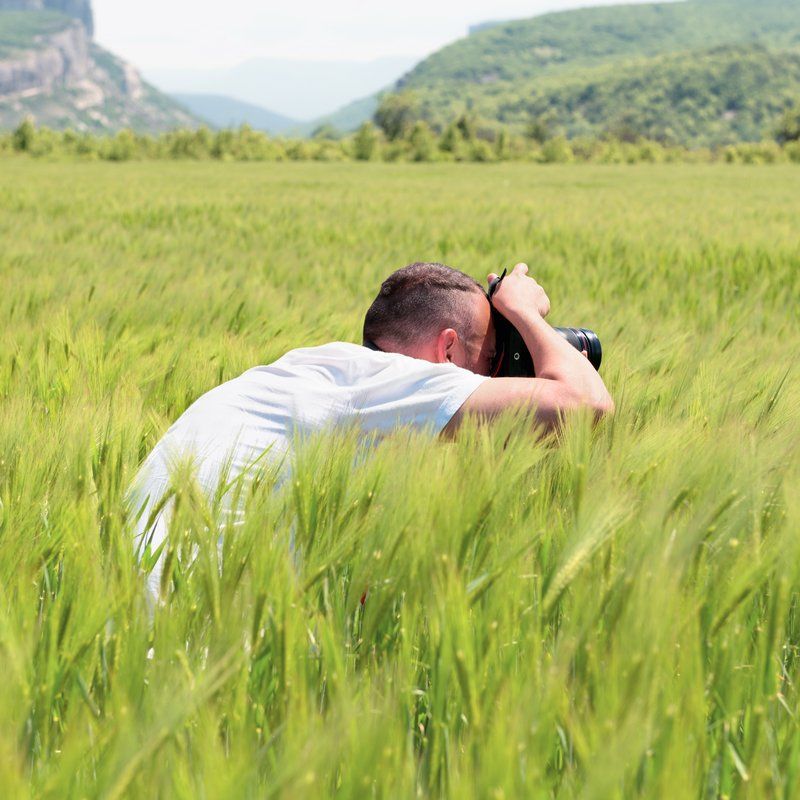 Photographer taking photo in tall green field with mountains in background.