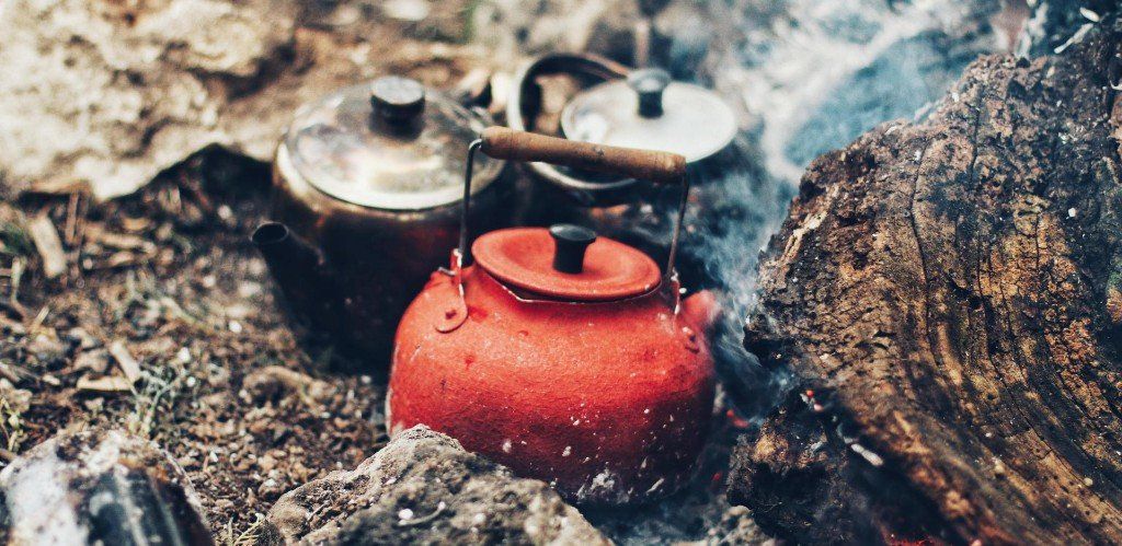 Red kettle boiling water over a campfire, with two other metal pots nearby.