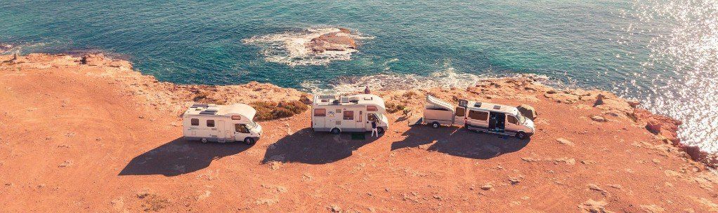 Three RVs parked on a red-brown coastline overlooking the ocean. The middle RV's door is open.