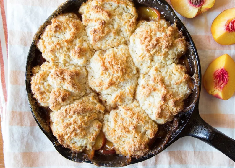 Peach cobbler in a cast iron skillet with golden biscuits, fresh peaches on a striped cloth.