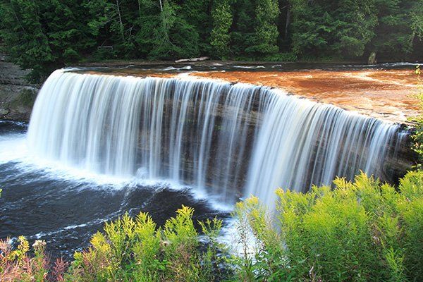 Waterfall cascading over a rocky cliff into a dark pool, surrounded by green trees and foliage.