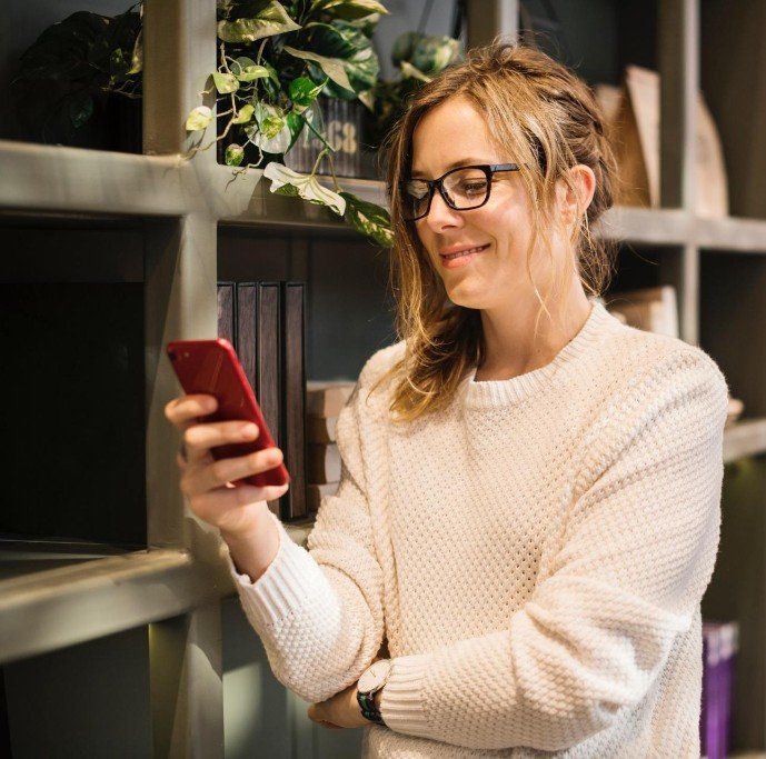 Woman wearing glasses looks at a red smartphone, smiling in front of a bookshelf.