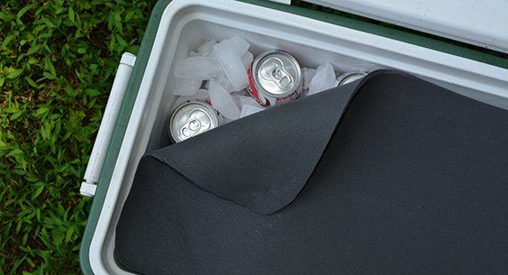 A cooler with ice and canned drinks, partially covered by a black mat, outdoors on grass.