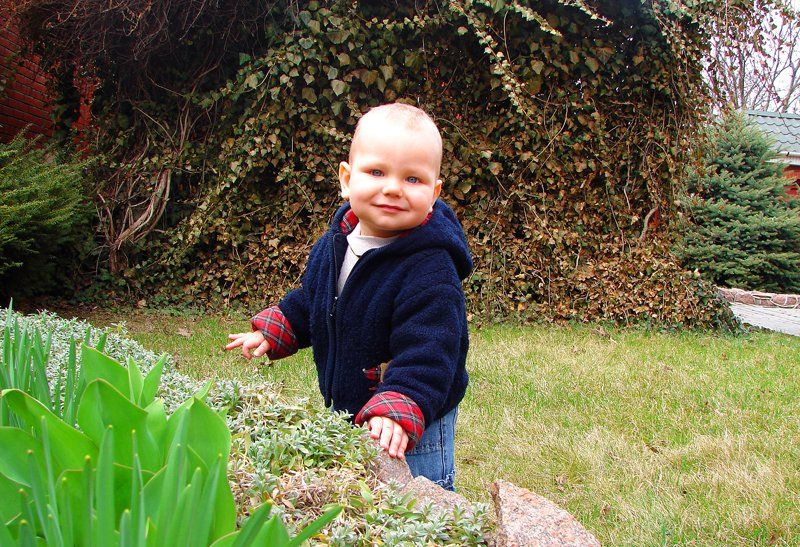 Baby standing in a garden, smiling, wearing a dark blue hooded jacket.