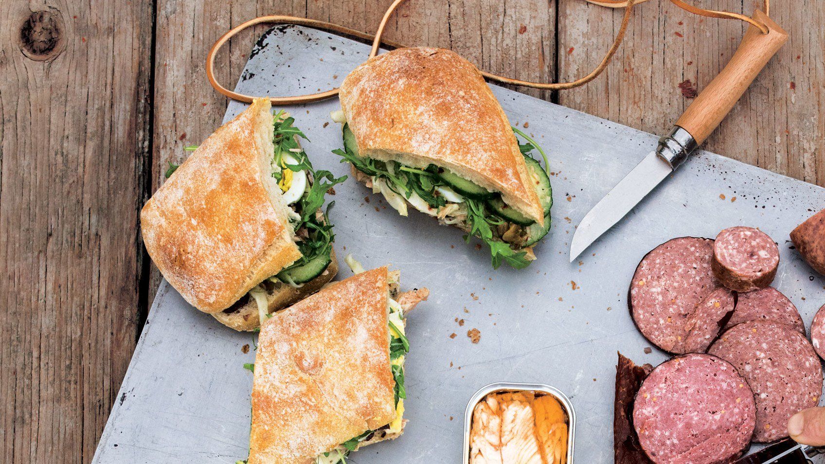 Sandwiches, sliced sausage, and tinned food on a gray platter and wooden surface.