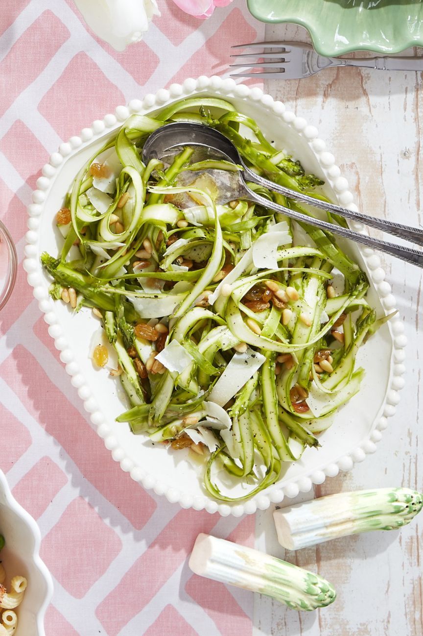 Asparagus salad in white dish on pink and white tablecloth with serving utensils and asparagus.