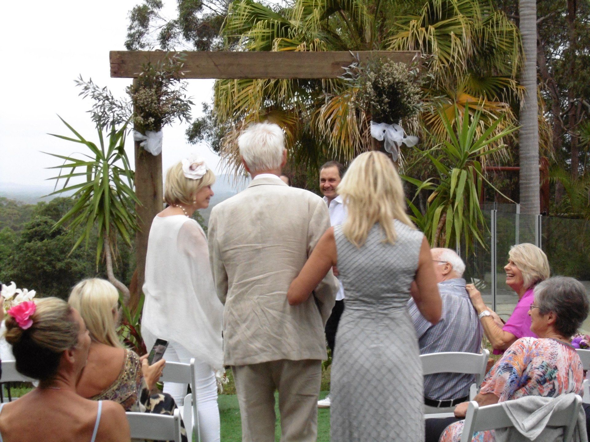 mum and dad in white and beige walking their daughter down aisle to groom as she gets married in garden at home