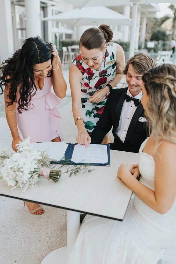 young bridal couple sit at table signing papers as mums are witnesses, celebrant in red points where to sign
