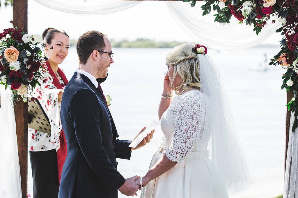 bride in white wiping happy tears as groom in navy suit reads his wedding vows to her, celebrant in red holding microphone