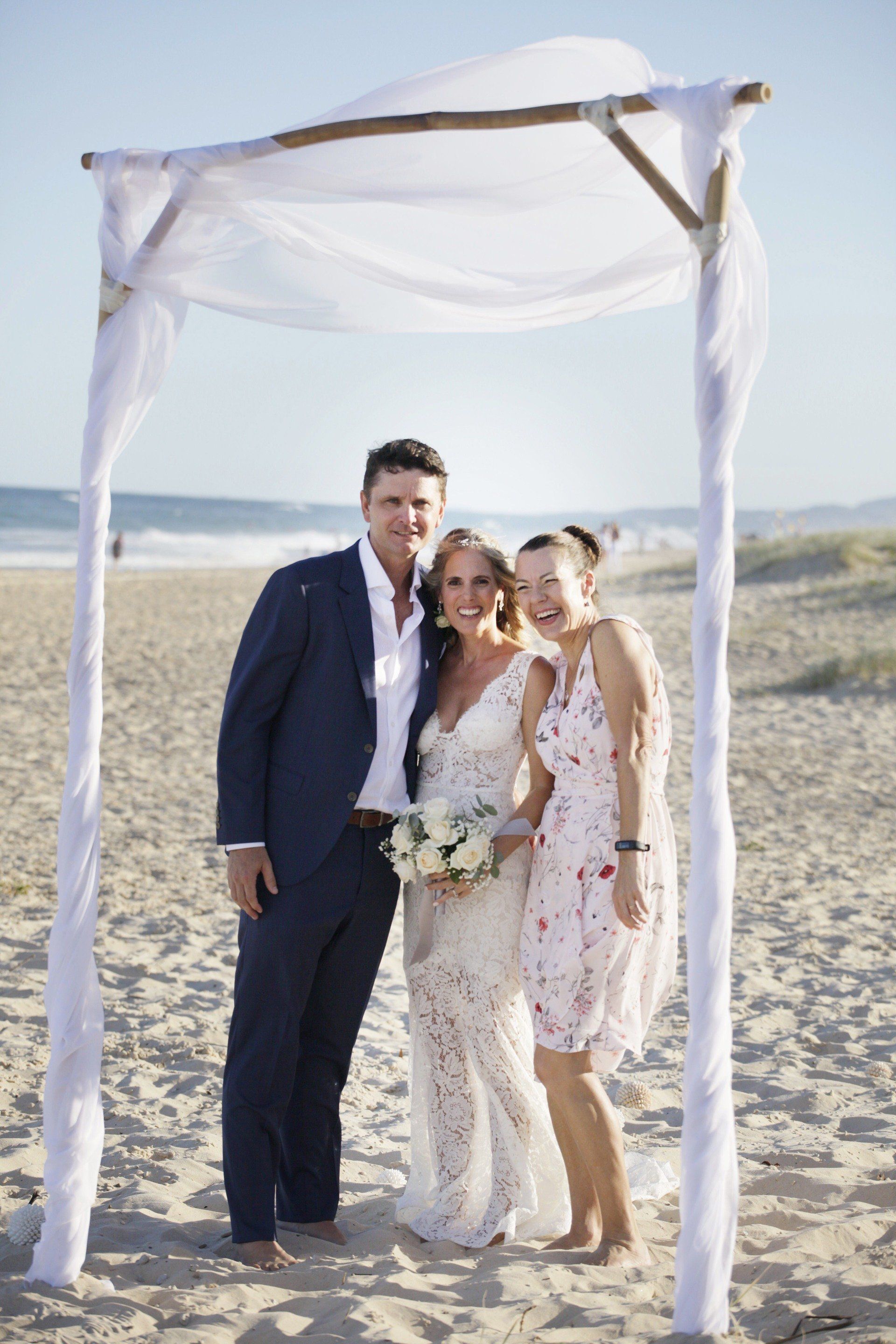 bride and groom and laughing celebrant on Sunshine beach with arbor and ocean on sunny day