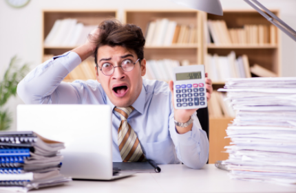 Man at desk looks shocked while holding a calculator, with laptop and stacks of papers in an office