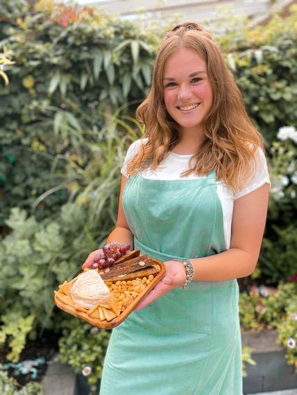 Een glimlachend persoon met een groen schort houdt een houten dienblad vast met crackers, kaas, noten en druiven in een tuinomgeving.