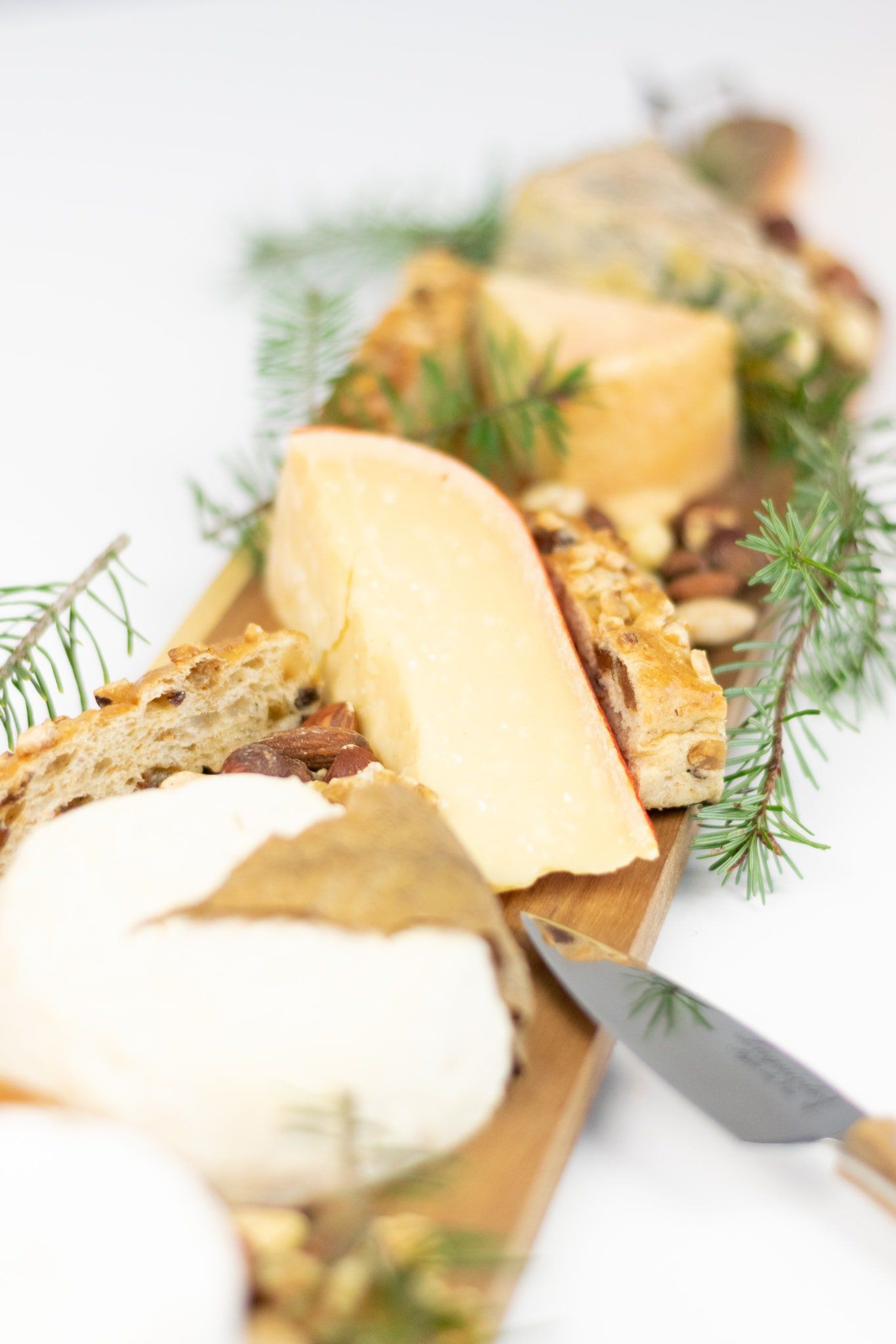 Op een houten plank liggen diverse luxe kazen, knapperig brood en noten uitgestald, aangevuld met verse dennentakjes.