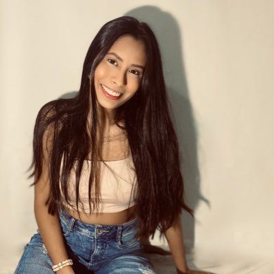 Una mujer con cabello largo está sentada en una cama vistiendo un top corto y jeans.