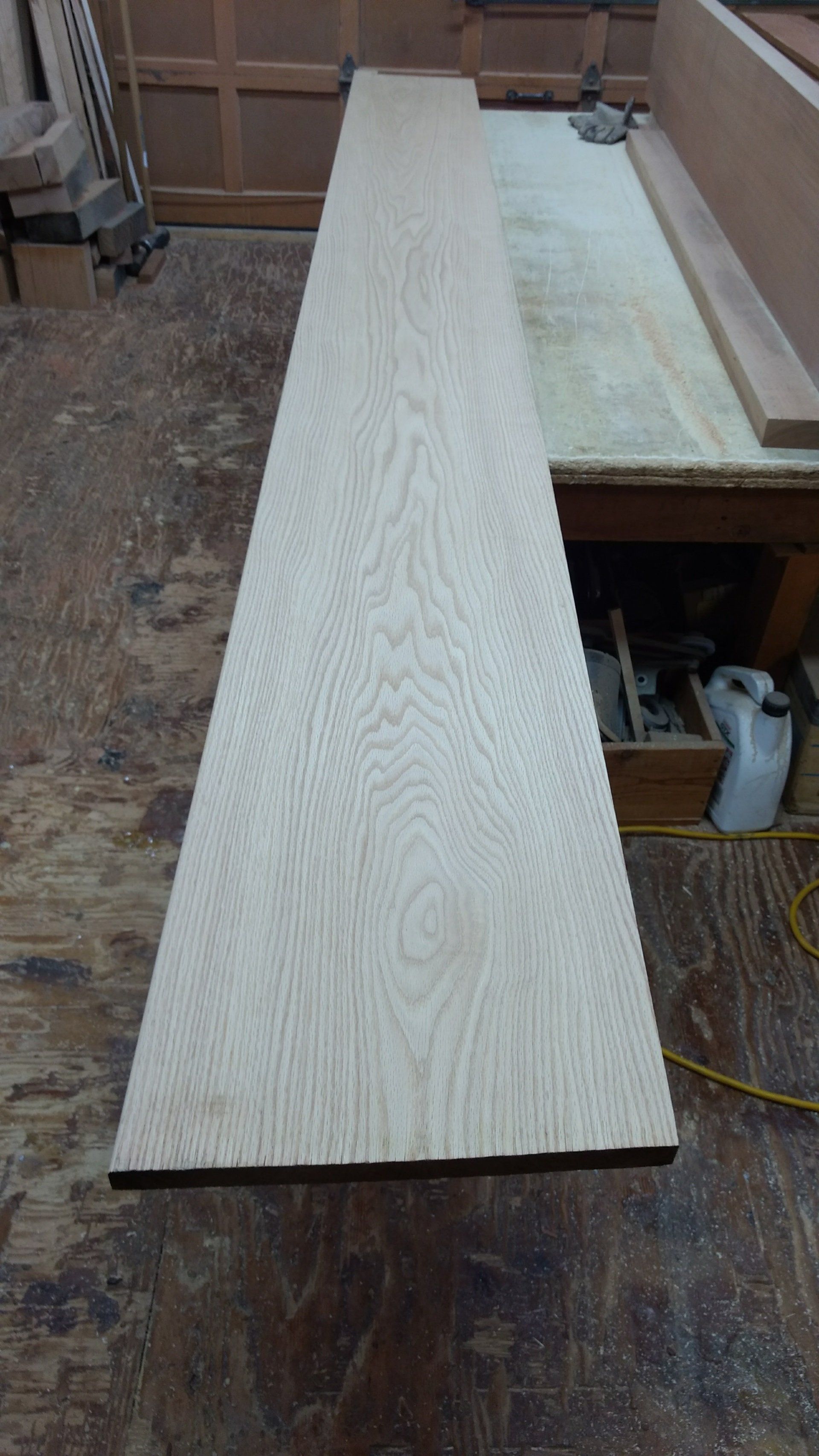 Long, light-colored wooden board on a workshop floor, showing wood grain.