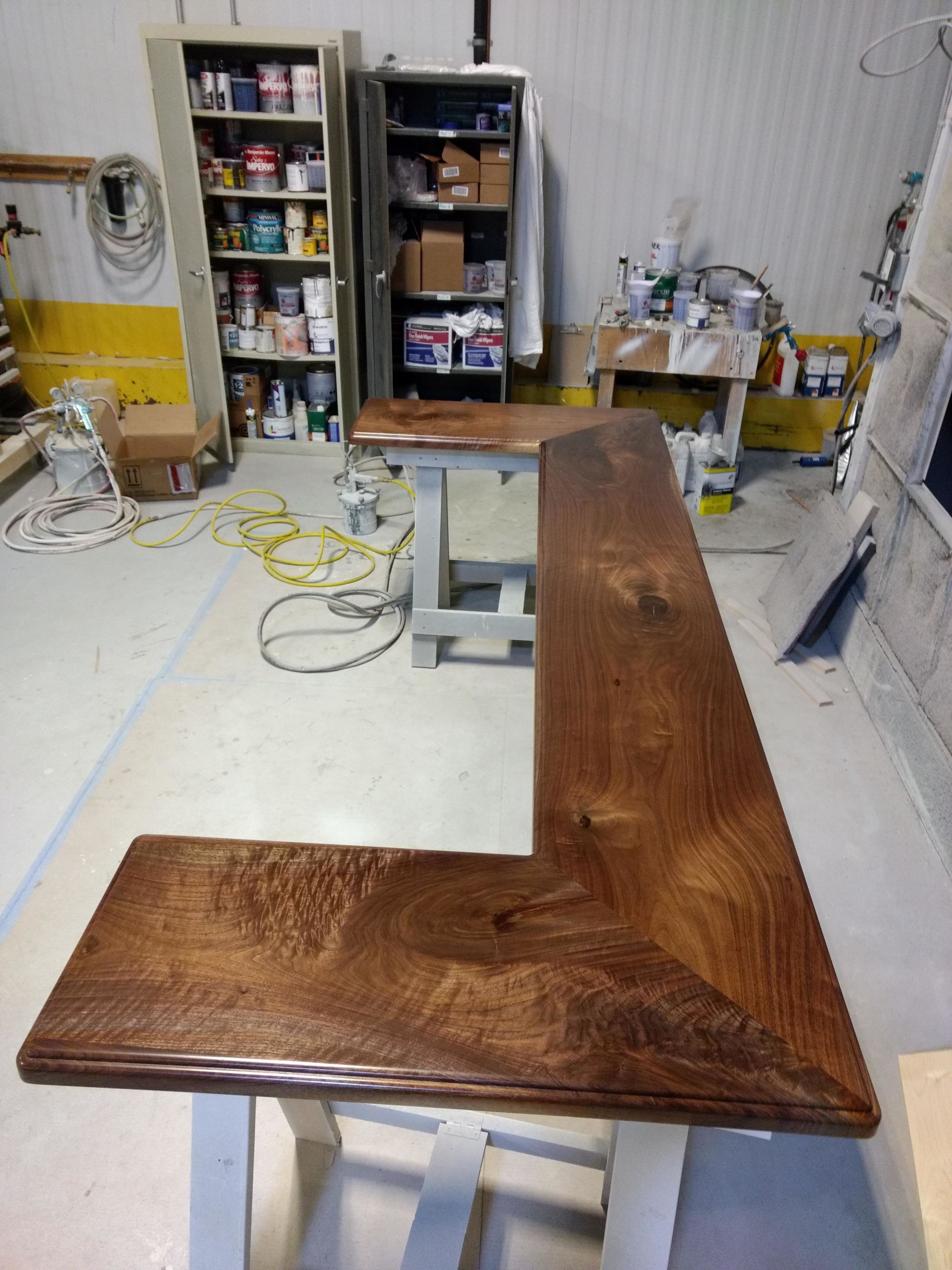L-shaped, stained wooden bar countertop on sawhorses in a workshop. Workshop contains shelving and supplies.