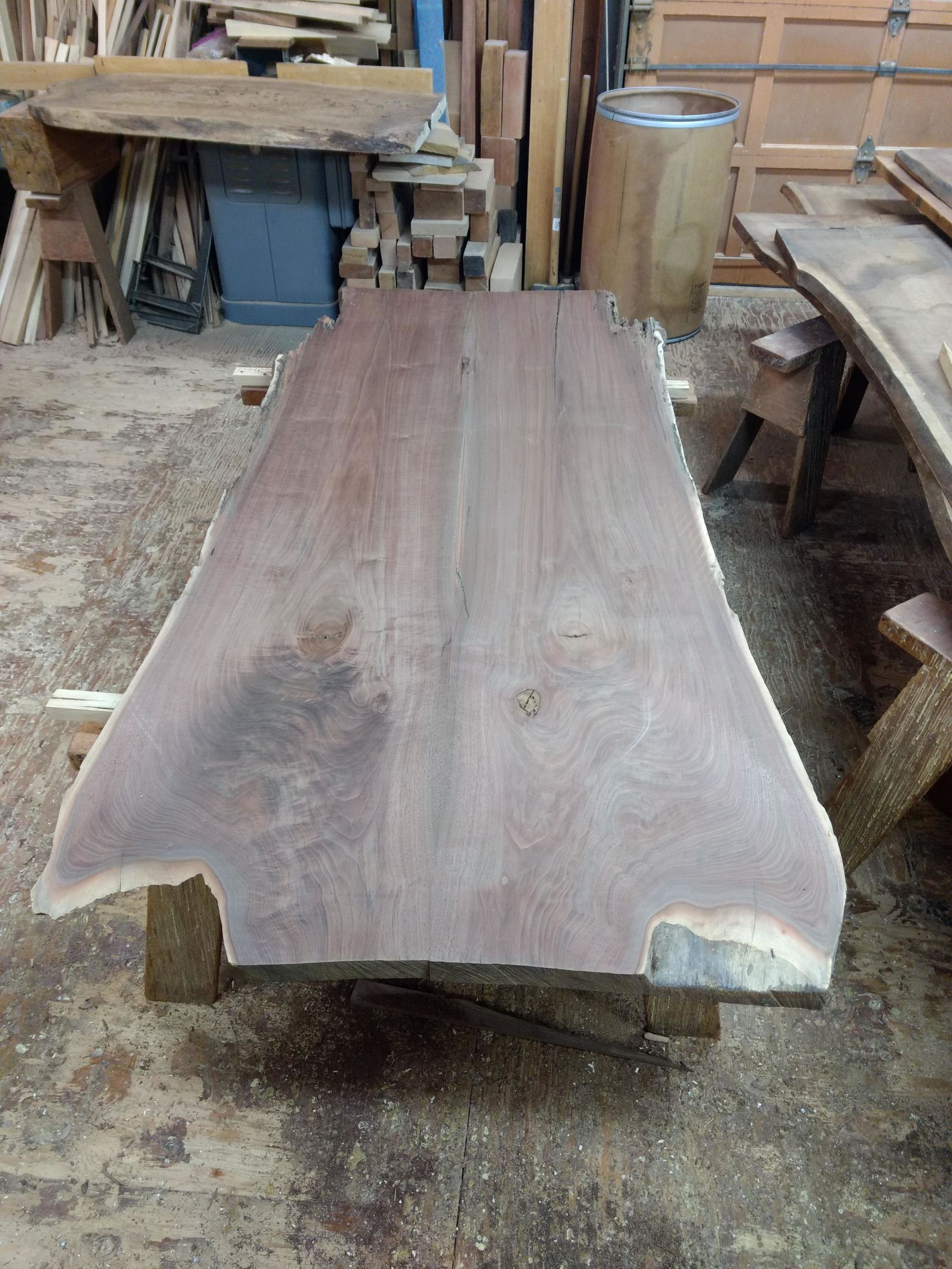 Live-edge wooden table in workshop, resting on supports, unfinished, brown wood.
