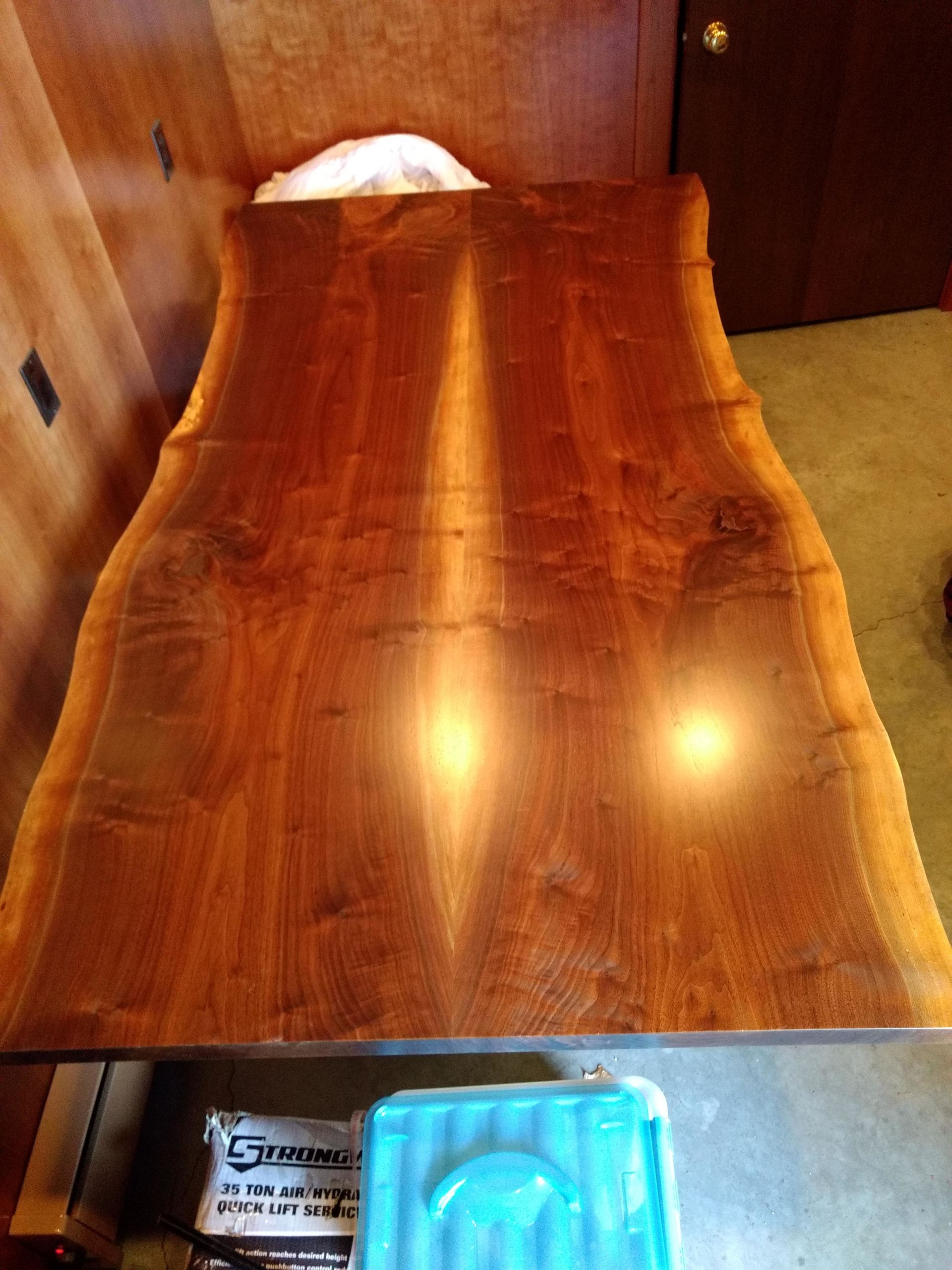 A large, rectangular wooden table with a natural edge, reflecting light. Turquoise container sits below it.