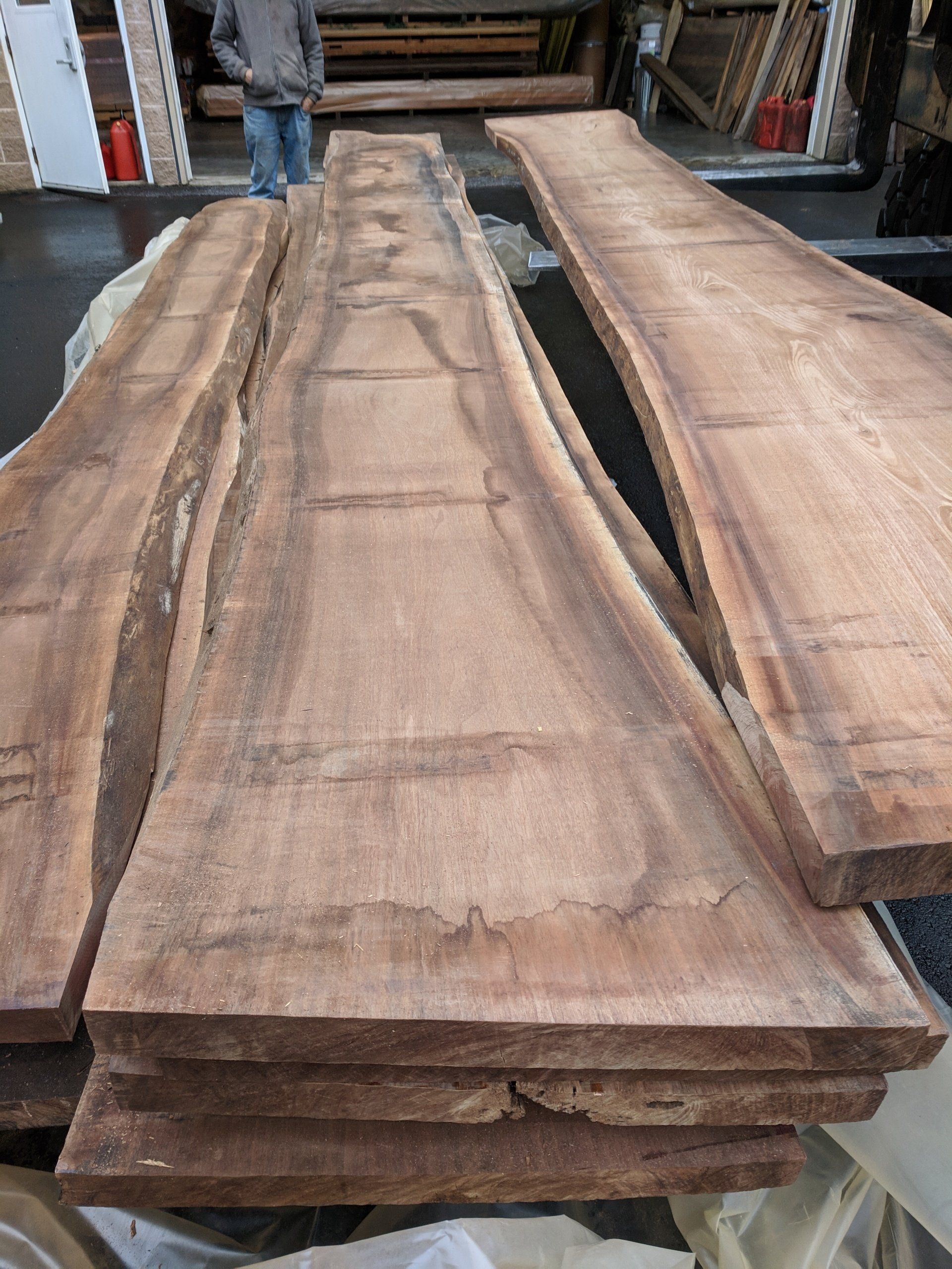 Stack of rough-cut, brown wood planks with live edges. A person stands in the background near a lumberyard.