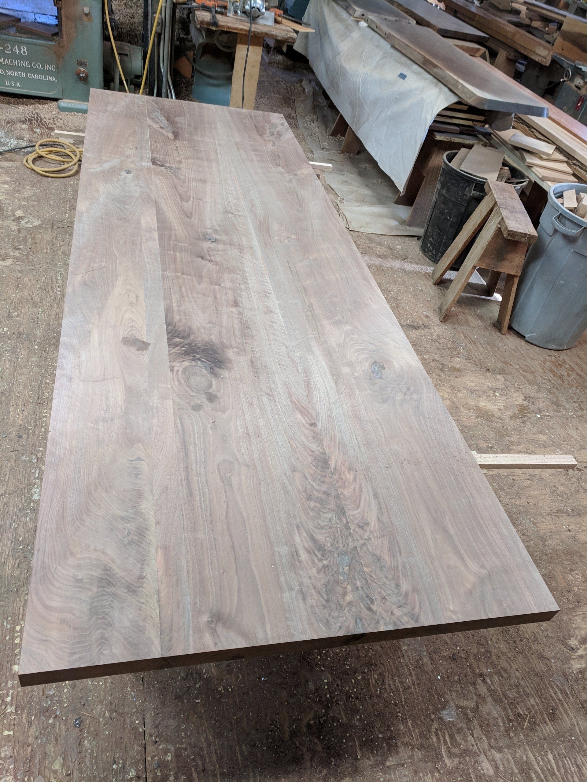 A large, rectangular wood tabletop sits on a concrete floor in a workshop.