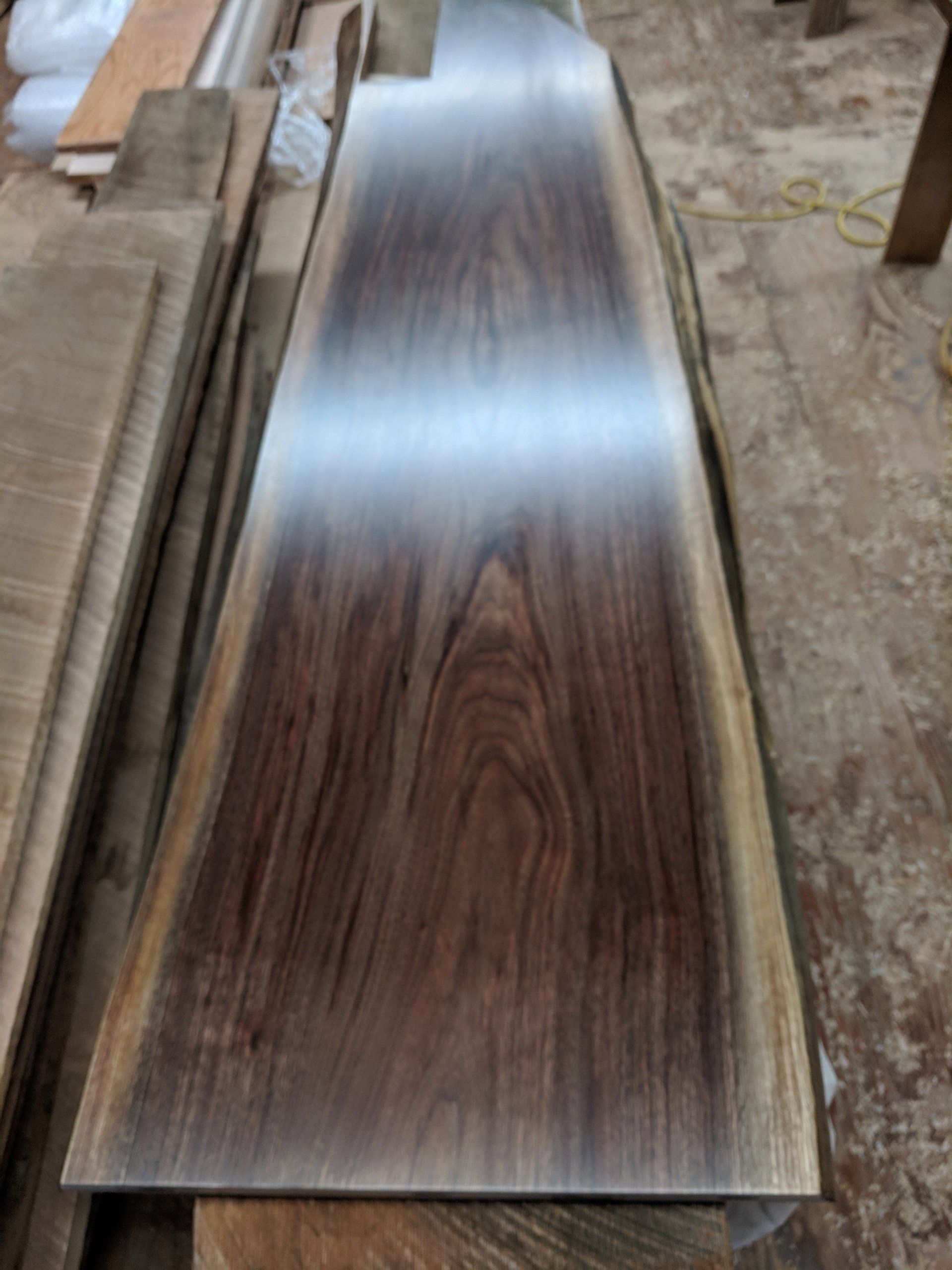 A long, dark-grained wooden slab with a glossy finish. It's set on a workshop floor, showing its natural edge.