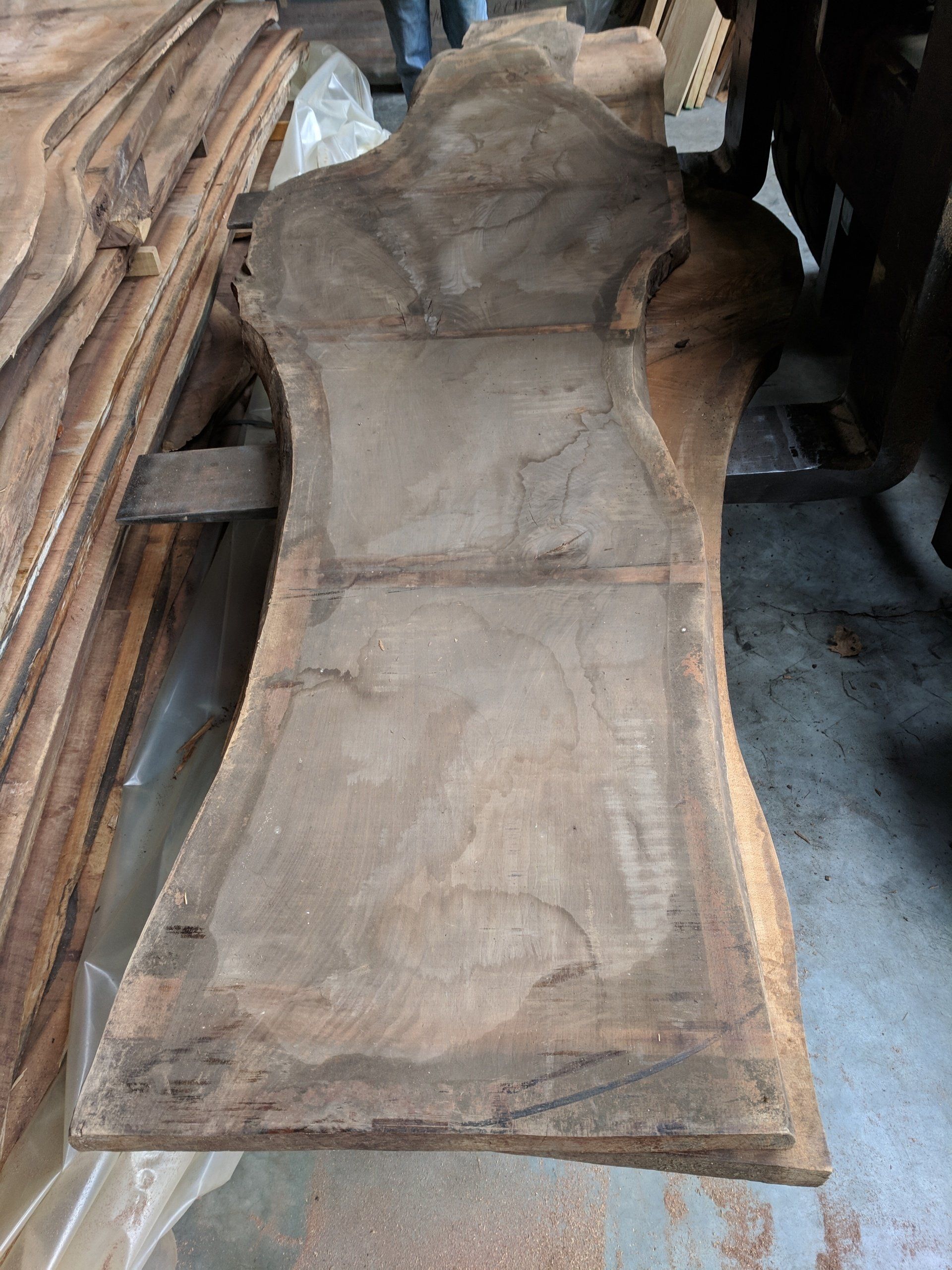 Large, rough-edged slab of dark wood, possibly walnut, with a natural, irregular shape, stacked with other boards.