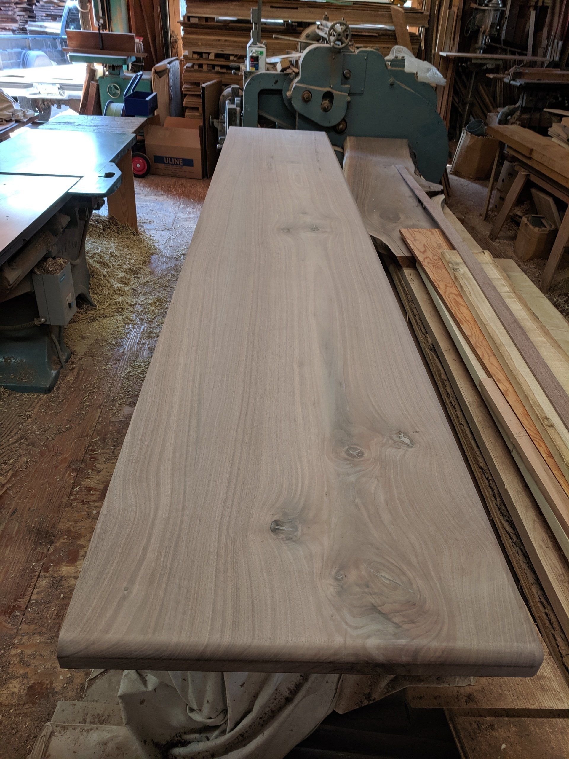 Wooden tabletop in a workshop.  Rounded edge, natural wood grain. Other woodworking equipment is in the background.