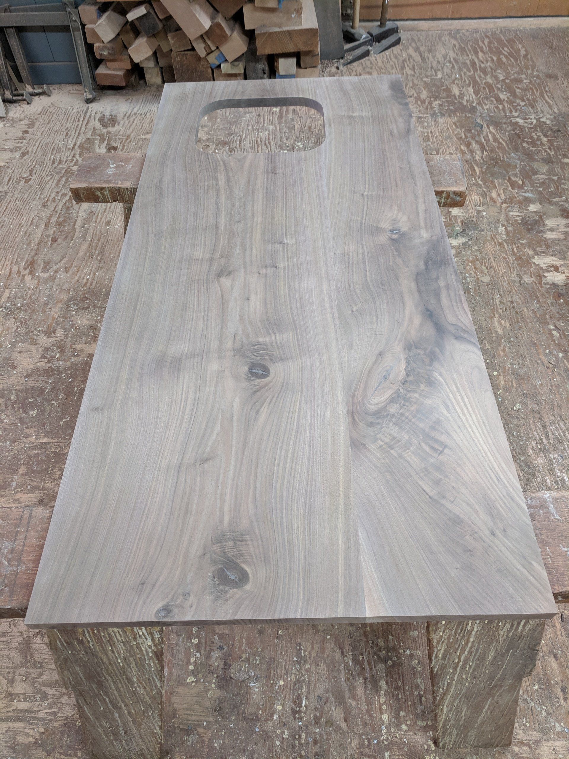 Wooden table with rectangular handle cutout, stained gray, resting on wooden supports.