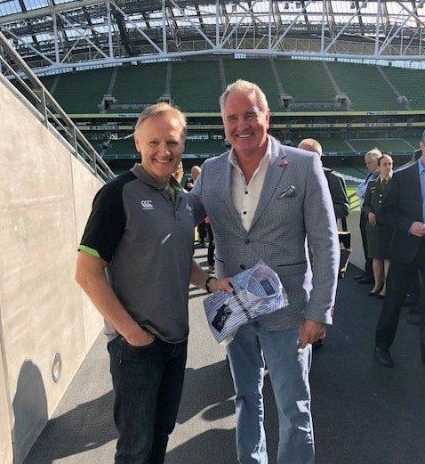 Two men standing side-by-side inside a stadium. One wears a green-accented shirt, the other a light blue jacket and jeans.