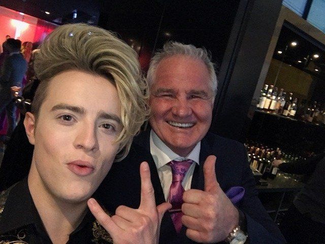 Man with blonde hair makes a selfie with older man, thumbs up, in a bar setting with bottles in background.