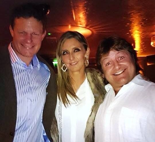 Three smiling people indoors, woman centered, flanked by two men, dim lighting.
