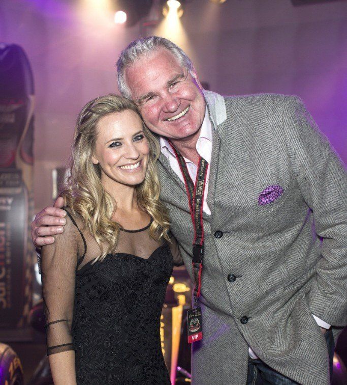 A man in a gray jacket and a woman in a black dress smile, posing together at an event with purple lighting.