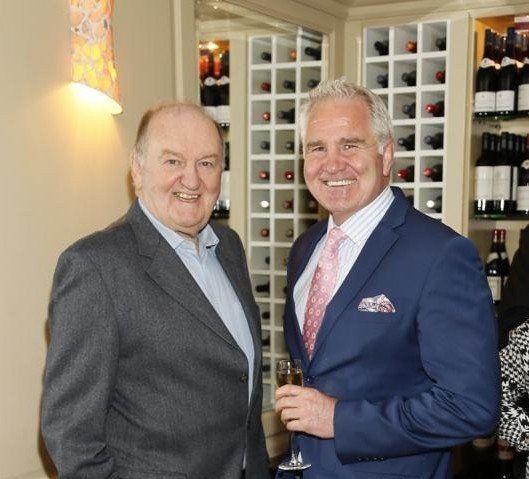 Two men smiling, one in a suit with a pink tie, the other in a blazer, standing near a wine rack.