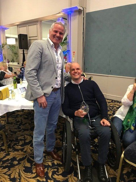 Man in a wheelchair smiles with a man in a blazer; they stand near a table in a room.